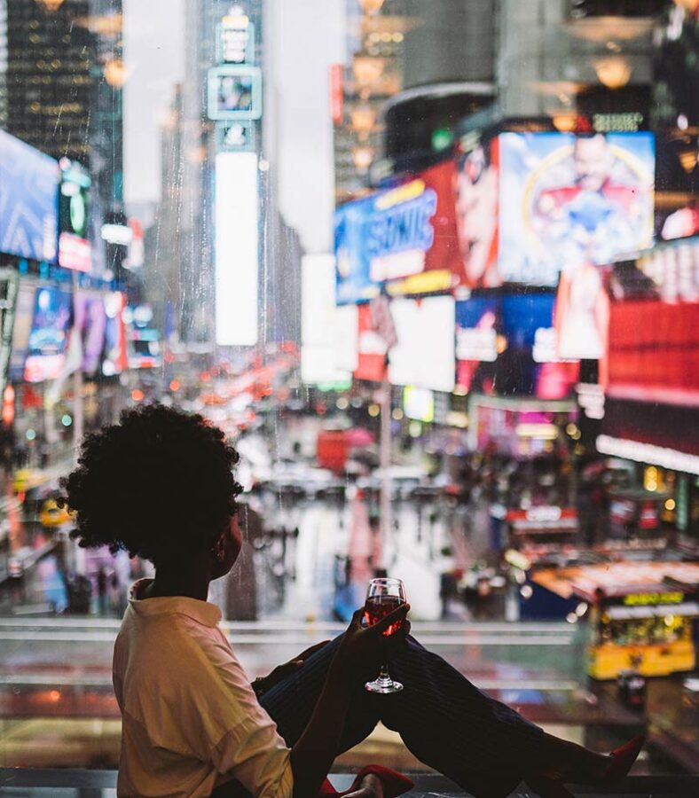 a person holding a glass of wine and sitting in front of windows looking out over Times Square