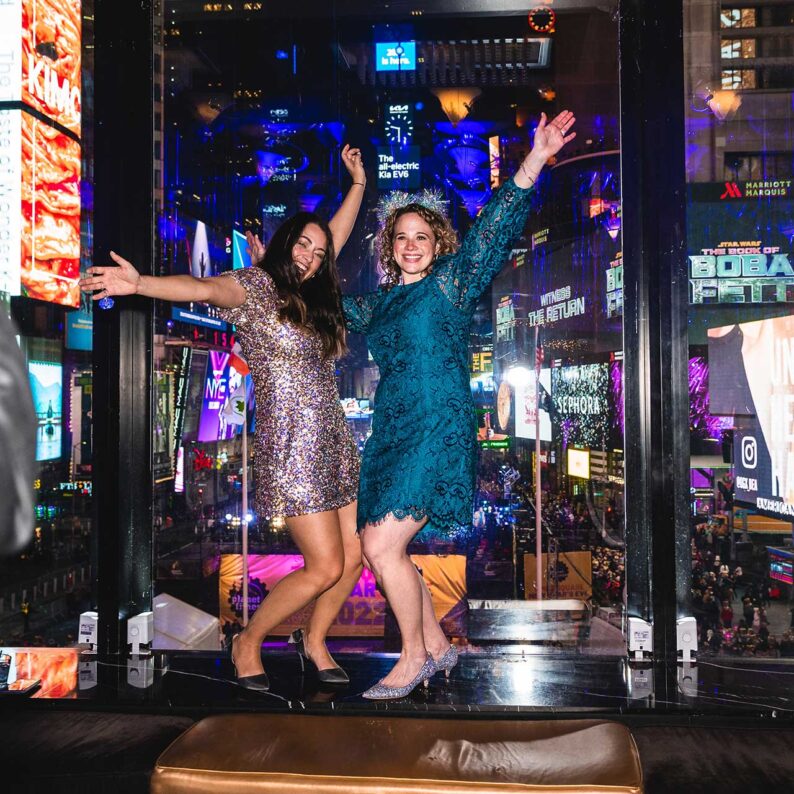 two smiling women in dresses with arms raised in front of window overlooking times square